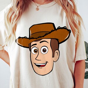 May include: Cream-colored t-shirt featuring a cartoon illustration of a character wearing a brown cowboy hat. The character has brown hair, a smiling face, and two different colored eyes. The design is outlined in black.