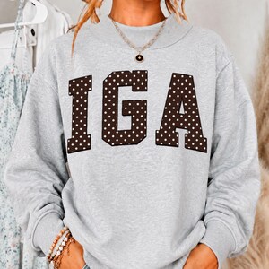 May include: A light gray long-sleeve sweatshirt featuring the letters IGA in large, dark brown block font with a white polka dot pattern. The garment has a crew neckline and is paired with a thin gold chain necklace featuring a small circular pendant.