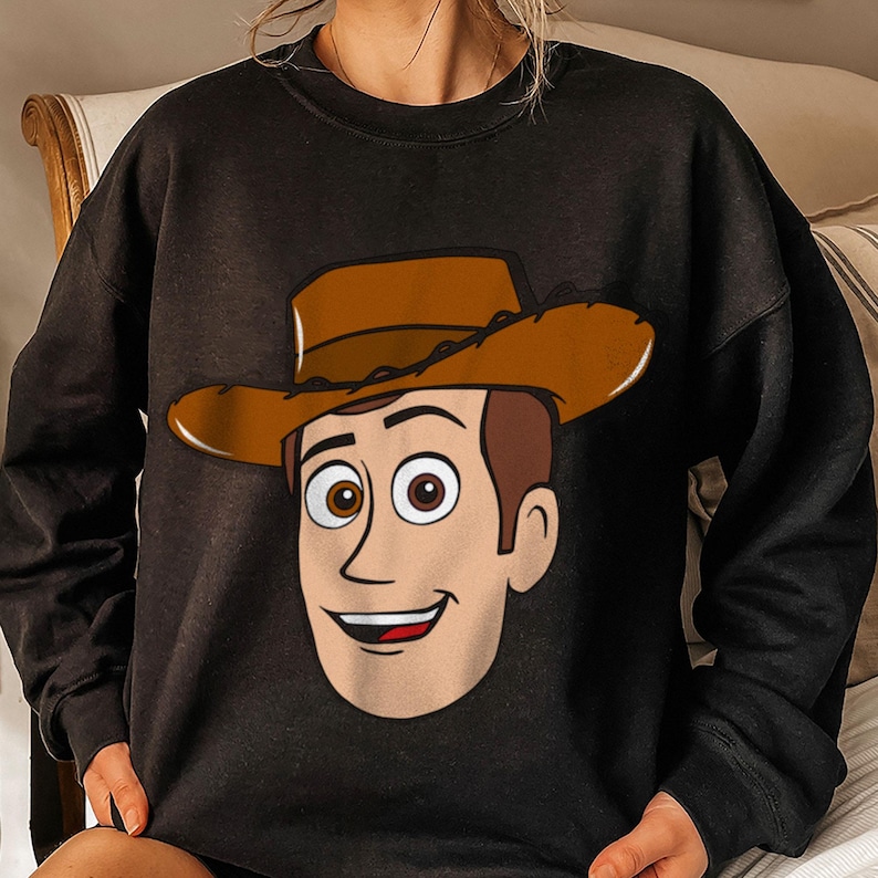 May include: Black sweatshirt featuring a cartoon illustration of a cowboy character's face. The character has brown hair, a brown cowboy hat, and a smiling expression. The sweatshirt is a solid black color.