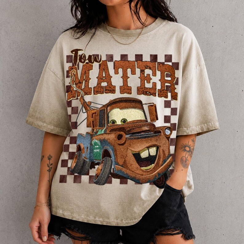 May include: A beige short-sleeve t-shirt featuring a brown and white checkered background pattern. The graphic displays a rusty brown tow truck character with large eyes and a wide smile, accompanied by the text Tow Mater in brown lettering.