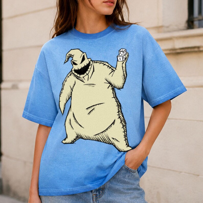 May include: A light blue short-sleeve t-shirt featuring a large graphic print of a beige, sack-like character with a stitched appearance, a wide jagged grin, and dark eyes, holding two white dice in one hand against the blue fabric background.