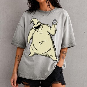 May include: A grey acid-wash t-shirt featuring a large graphic print of a pale yellow, sack-like character with a wide, dark, jagged mouth. The character is depicted holding a pair of white dice in one hand, with visible black stitching details.