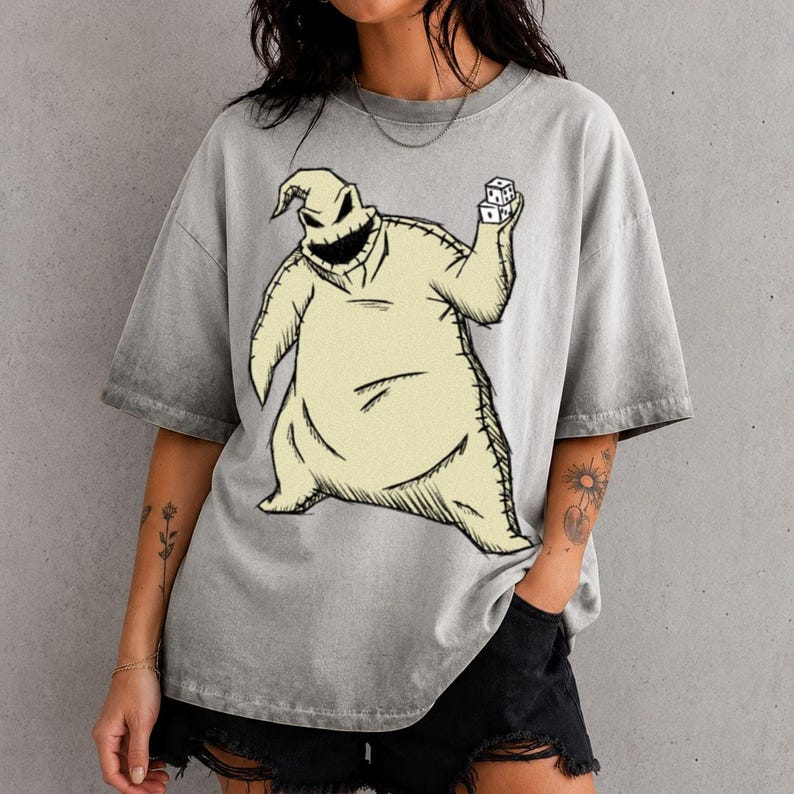 May include: A grey acid-wash t-shirt featuring a large graphic print of a pale yellow, sack-like character with a wide, dark, jagged mouth. The character is depicted holding a pair of white dice in one hand, with visible black stitching details.