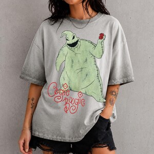 May include: A grey acid-wash t-shirt featuring a graphic of the character Oogie Boogie from The Nightmare Before Christmas. The character is light green with stitched details, holding red dice. The text "Oogie Boogie" is printed in red below.