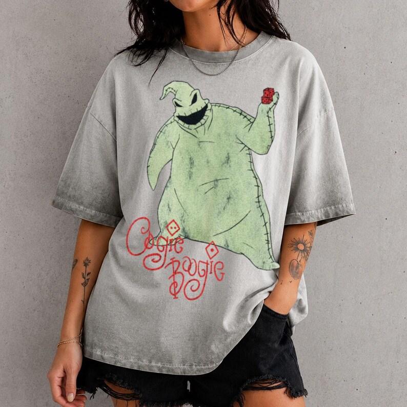 May include: A grey acid-wash t-shirt featuring a graphic of the character Oogie Boogie from The Nightmare Before Christmas. The character is light green with stitched details, holding red dice. The text "Oogie Boogie" is printed in red below.