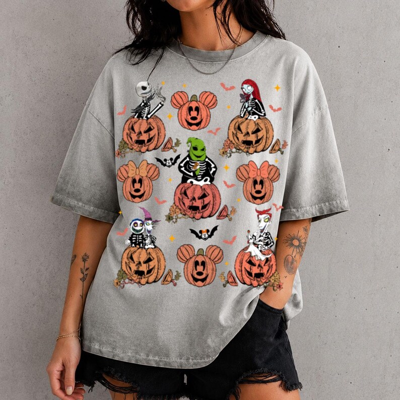 May include: A grey acid-wash t-shirt featuring a graphic print of various characters from The Nightmare Before Christmas sitting in or on orange pumpkins, interspersed with orange Mickey Mouse-shaped pumpkins, small bats, and orange stars.