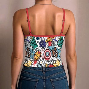K&ouml;nnte beinhalten: Wei&szlig;es Camisole mit einem farbenfrohen Comic-Design mit verschiedenen Superhelden-Symbolen und -Charakteren. Das Camisole hat rote Tr&auml;ger und Bes&auml;tze und wird mit einer blauen Jeans getragen.