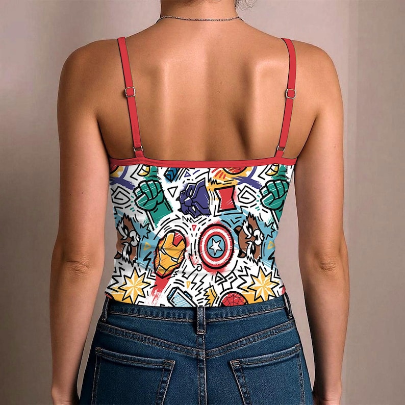 K&ouml;nnte beinhalten: Wei&szlig;es Camisole mit einem farbenfrohen Comic-Design mit verschiedenen Superhelden-Symbolen und -Charakteren. Das Camisole hat rote Tr&auml;ger und Bes&auml;tze und wird mit einer blauen Jeans getragen.
