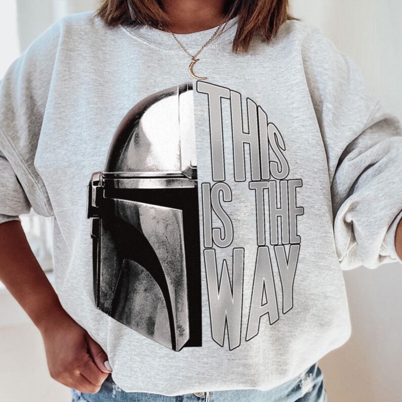 May include: A light gray sweatshirt featuring a graphic of a Mandalorian helmet and the text "THIS IS THE WAY" in gray. The design is centered on the front of the long-sleeved crew neck top.