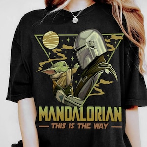 May include: Black t-shirt featuring a graphic of The Mandalorian and Grogu. The design includes a helmeted figure and a small green creature, set against a triangle with a planet and stars. The text "MANDALORIAN - THIS IS THE WAY" is printed below.