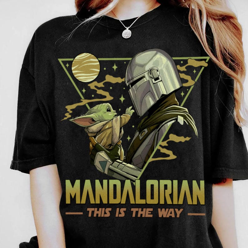 May include: Black t-shirt featuring a graphic of The Mandalorian and Grogu. The design includes a helmeted figure and a small green creature, set against a triangle with a planet and stars. The text "MANDALORIAN - THIS IS THE WAY" is printed below.