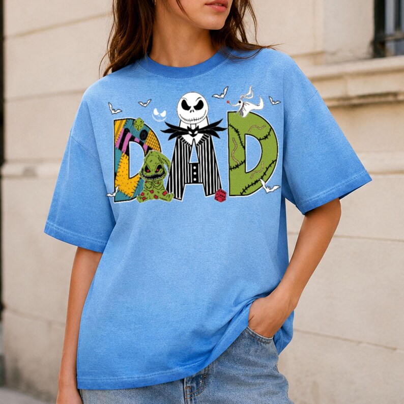 May include: A light blue t-shirt featuring the word DAD in large letters. The letter D is a yellow and purple patchwork pattern, the A is a black and white pinstriped suit with a skull head, and the D is green with stitches and a ghost dog.