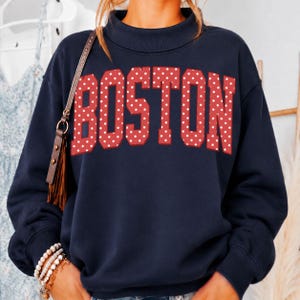 May include: A navy blue long-sleeve sweatshirt featuring a mock neck collar and the word BOSTON printed in large, bold, red block letters with a white polka dot pattern. The garment has ribbed cuffs and a ribbed hem, providing a structured fit.