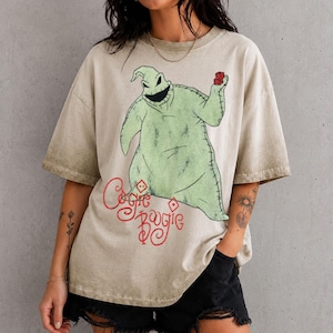 May include: A beige, acid-washed short-sleeve t-shirt featuring a large graphic of the character Oogie Boogie from The Nightmare Before Christmas. The character is light green with stitched details, holding red dice. The text Oogie Boogie is in red.