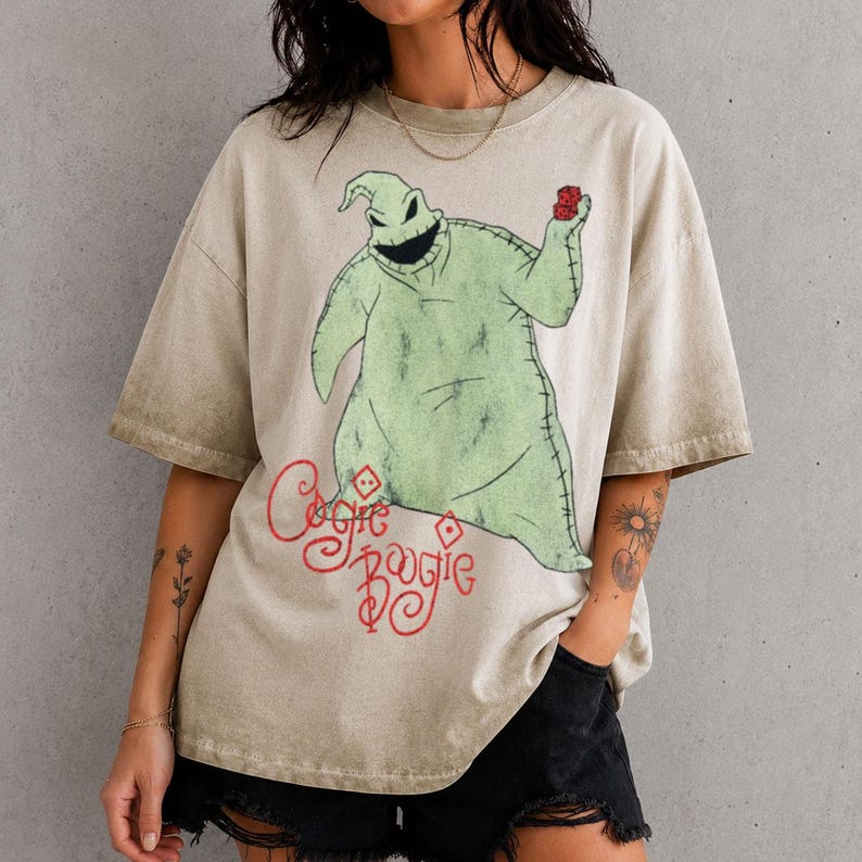 May include: A beige, acid-washed short-sleeve t-shirt featuring a large graphic of the character Oogie Boogie from The Nightmare Before Christmas. The character is light green with stitched details, holding red dice. The text Oogie Boogie is in red.