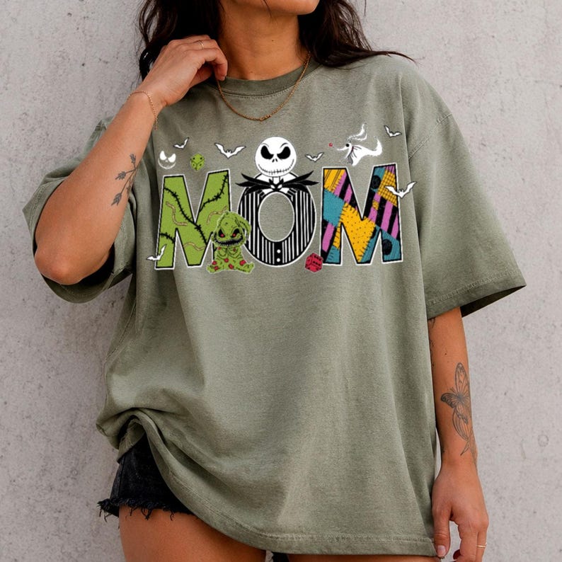 Puede incluir: Una camiseta verde oliva con la palabra MOM en letras grandes. La M es verde con puntadas, la O es la cara de Jack Skellington y la M es un patr&oacute;n de patchwork. Una peque&ntilde;a criatura verde y un fantasma blanco est&aacute;n impresos cerca de las letras.