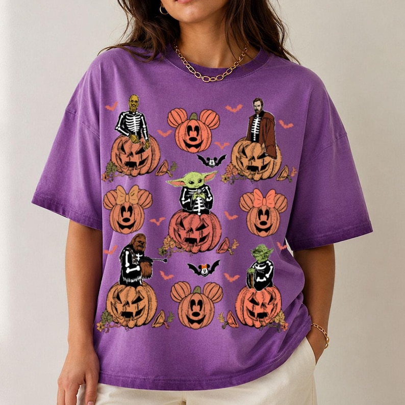 May include: A purple short-sleeve t-shirt featuring a graphic print of various characters wearing skeleton costumes, sitting on orange jack-o'-lanterns. The design includes Mickey Mouse-shaped pumpkins, bats, and small orange floral accents.