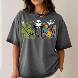 Puede incluir: Una camiseta de manga corta gris carb&oacute;n con la palabra MOM en letras grandes. La M es verde con puntadas, la O es una cara de Jack Skellington a rayas blancas y negras, y la M es un patr&oacute;n de patchwork colorido con murci&eacute;lagos y dados.