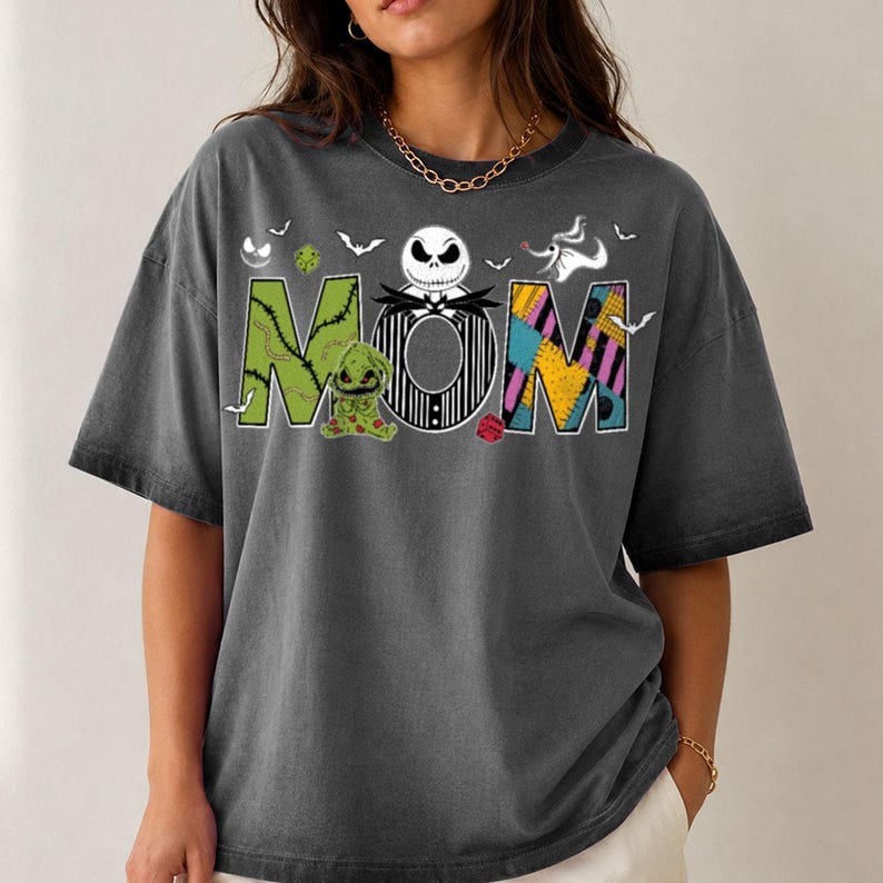Puede incluir: Una camiseta de manga corta gris carb&oacute;n con la palabra MOM en letras grandes. La M es verde con puntadas, la O es una cara de Jack Skellington a rayas blancas y negras, y la M es un patr&oacute;n de patchwork colorido con murci&eacute;lagos y dados.