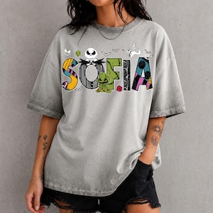 May include: A grey acid-wash short-sleeve t-shirt featuring the name SOFIA in large, colorful, patterned letters. The design includes a skull character, a green creature, and small bat silhouettes inspired by The Nightmare Before Christmas.