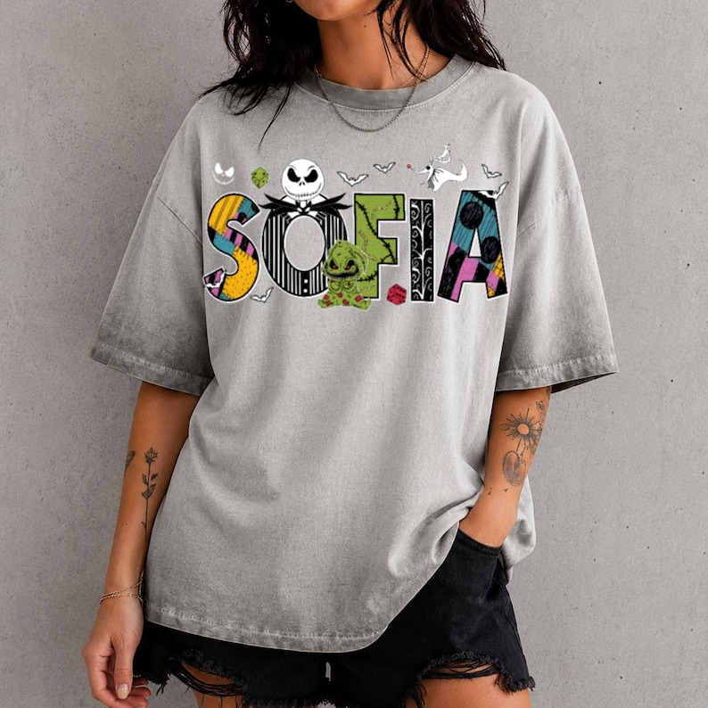 May include: A grey acid-wash short-sleeve t-shirt featuring the name SOFIA in large, colorful, patterned letters. The design includes a skull character, a green creature, and small bat silhouettes inspired by The Nightmare Before Christmas.
