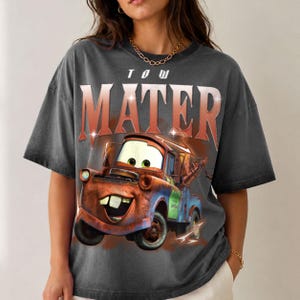 K&ouml;nnte beinhalten: Ein anthrazitfarbenes Kurzarm-T-Shirt mit einem gro&szlig;en Grafikdruck der Figur Tow Mater aus dem Film Cars. Der Text TOW MATER ist in einer fetten, abgenutzten roten Schrift &uuml;ber der Illustration des rostigen Abschleppwagens aufgedruckt.