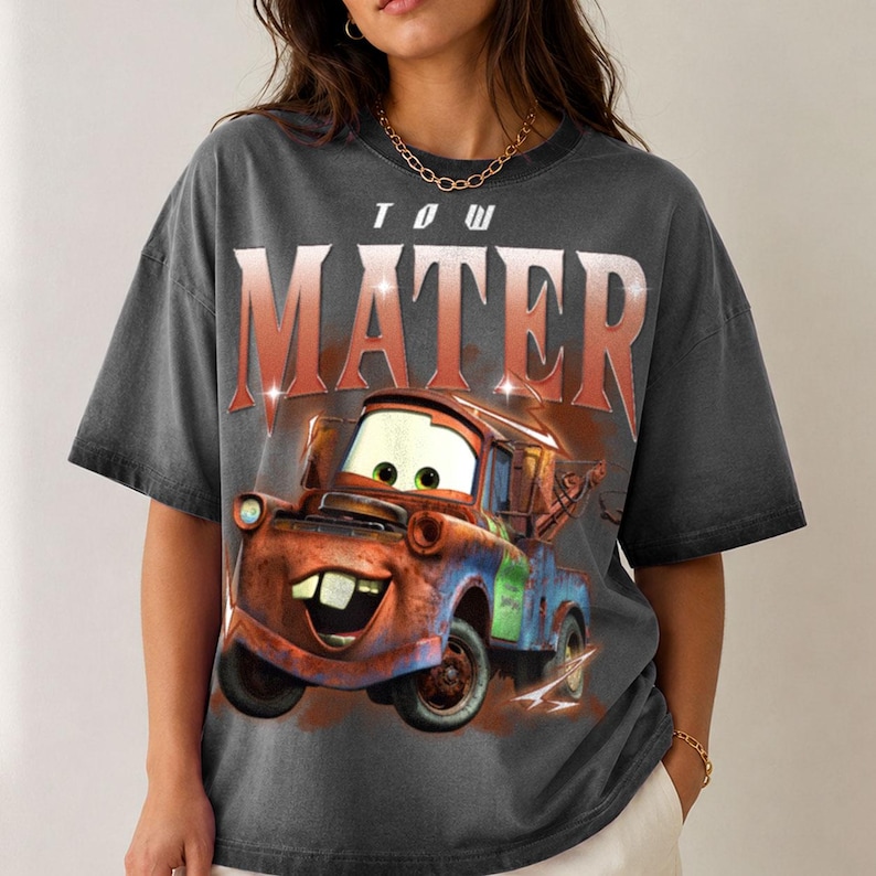 K&ouml;nnte beinhalten: Ein anthrazitfarbenes Kurzarm-T-Shirt mit einem gro&szlig;en Grafikdruck der Figur Tow Mater aus dem Film Cars. Der Text TOW MATER ist in einer fetten, abgenutzten roten Schrift &uuml;ber der Illustration des rostigen Abschleppwagens aufgedruckt.