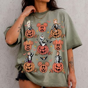以下が含まれることがあります： オリーブグリーンの半袖Tシャツ。骸骨の衣装を着た様々な漫画のキャラクターが、それぞれ彫られたジャック・オー・ランタンから顔を出しているグラフィックプリントが施されている。デザインにはミッキーの形をしたカボチャと小さなコウモリが含まれている。