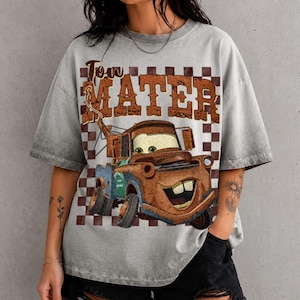 May include: A grey short-sleeve t-shirt featuring a graphic print of a rusty brown tow truck character with large eyes and a wide smile. Above the truck, the text "Tow Mater" is printed in a distressed brown font over a brown checkered background.