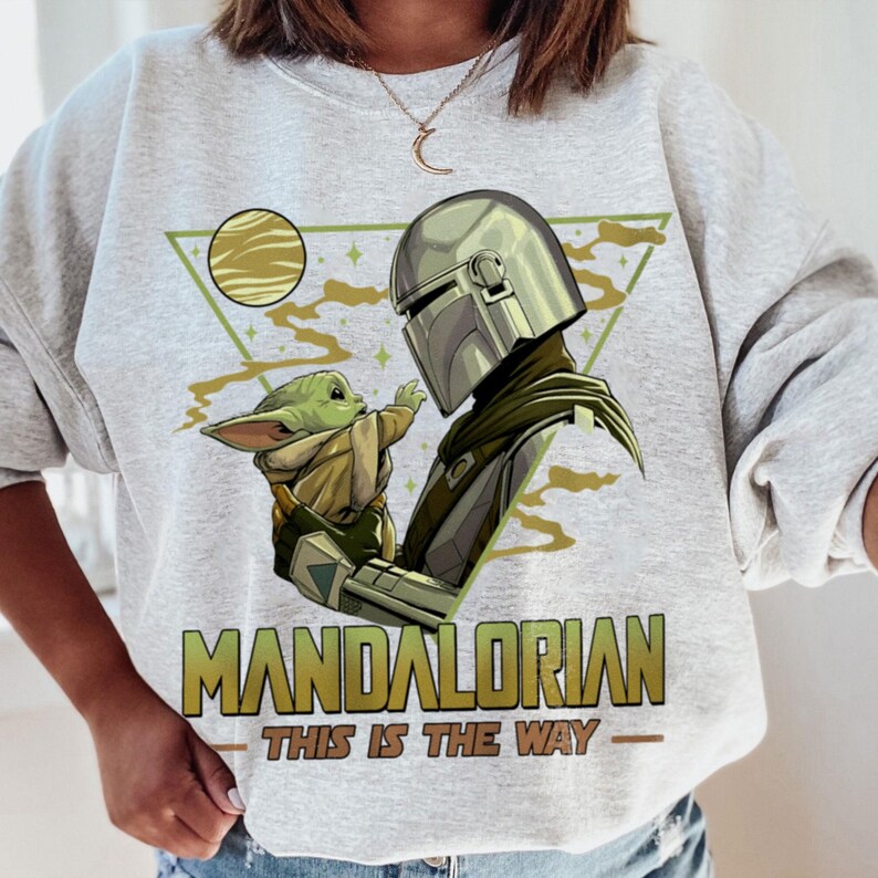 May include: A light gray sweatshirt featuring a graphic of The Mandalorian and Grogu. The design includes a green triangle, a yellow planet, and the text "MANDALORIAN - THIS IS THE WAY" in gold and white.