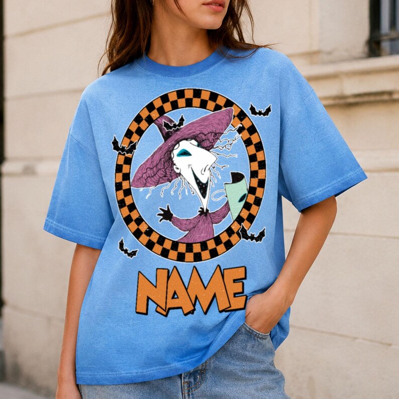 Puede incluir: Una camiseta de manga corta azul claro con un gr&aacute;fico de un personaje con sombrero y t&uacute;nica de bruja morada dentro de un c&iacute;rculo a cuadros negro y naranja. Peque&ntilde;os murci&eacute;lagos negros rodean el c&iacute;rculo y la palabra NAME est&aacute; impresa en naranja debajo.