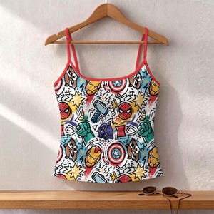 K&ouml;nnte beinhalten: Ein wei&szlig;es Camisole mit roter Bord&uuml;re und Tr&auml;gern, mit einem farbenfrohen Muster aus Comicfiguren und Symbolen. Das Design umfasst Bilder von Spider-Man, Captain America und Thor.