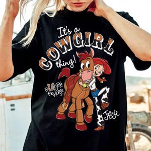 May include: Black t-shirt with a graphic featuring the text "It's a COWGIRL thing!" and cartoon characters Bullseye the Horse and Jessie. The design includes brown, white, red, and blue accents.
