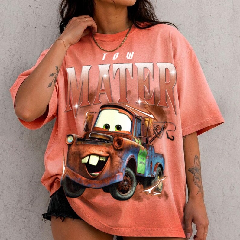 K&ouml;nnte beinhalten: Ein korallenfarbenes Kurzarm-T-Shirt mit einer gro&szlig;en Grafik der Figur Hook aus dem Film Cars. Der Text MATER ist in einer gro&szlig;en, metallisch wirkenden Schrift &uuml;ber der Abbildung des rostigen Abschleppwagens auf das T-Shirt gedruckt.