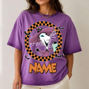 Puede incluir: Una camiseta morada de manga corta con un gr&aacute;fico de un personaje con sombrero y t&uacute;nica morados dentro de un c&iacute;rculo a cuadros negros y naranjas. El dise&ntilde;o incluye peque&ntilde;os murci&eacute;lagos negros y la palabra NAME en letras naranjas grandes.