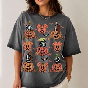 May include: A charcoal gray short-sleeve t-shirt featuring a Halloween-themed graphic print. The design includes various orange jack-o'-lanterns with mouse-ear shapes, small bats, and several characters wearing skeleton costumes sitting on pumpkins.