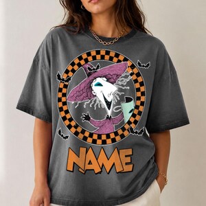 Puede incluir: Una camiseta gris carb&oacute;n con un gr&aacute;fico de un personaje con sombrero y t&uacute;nica de bruja morados, sosteniendo una m&aacute;scara. El personaje est&aacute; enmarcado por un c&iacute;rculo a cuadros naranja y negro con murci&eacute;lagos. La palabra NAME aparece en naranja debajo.