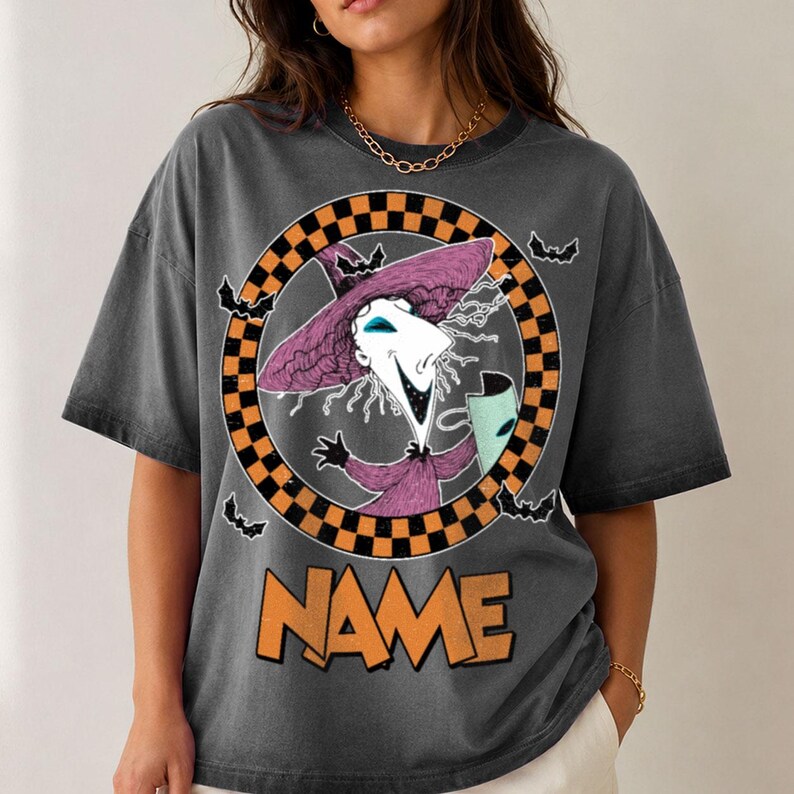 Puede incluir: Una camiseta gris carb&oacute;n con un gr&aacute;fico de un personaje con sombrero y t&uacute;nica de bruja morados, sosteniendo una m&aacute;scara. El personaje est&aacute; enmarcado por un c&iacute;rculo a cuadros naranja y negro con murci&eacute;lagos. La palabra NAME aparece en naranja debajo.