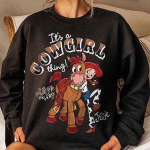 May include: Black sweatshirt featuring the phrase "It's a Cowgirl thing!" with an illustration of Jessie and Bullseye the horse. Jessie is wearing a cowgirl hat and outfit. Bullseye has a saddle. The sweatshirt is a comfortable, casual style.