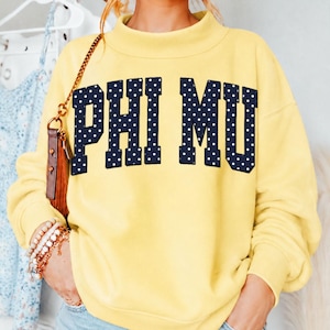 May include: A pale yellow long-sleeve sweatshirt with a mock neck collar. The front features the text PHI MU in large, dark navy blue block letters with a white polka dot pattern. The garment has ribbed cuffs and a matching ribbed hemline.