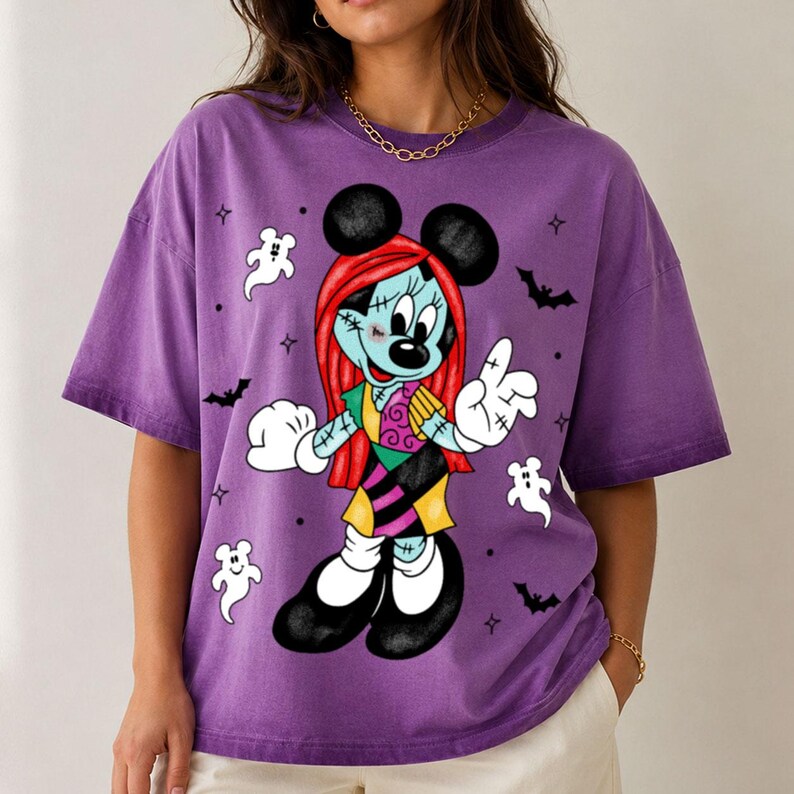 May include: A purple short-sleeve t-shirt featuring a graphic of a character with red hair, blue skin, and stitched details, wearing a patchwork dress. The design includes white ghost figures, black bats, and small star shapes scattered around.