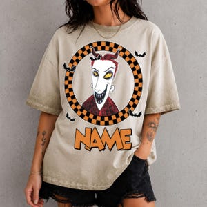 May include: A beige short-sleeve t-shirt featuring a graphic of a character with red hair, yellow eyes, and a wide grin inside a black and orange checkered circle. Small black bats surround the circle, and the word NAME is printed in orange.
