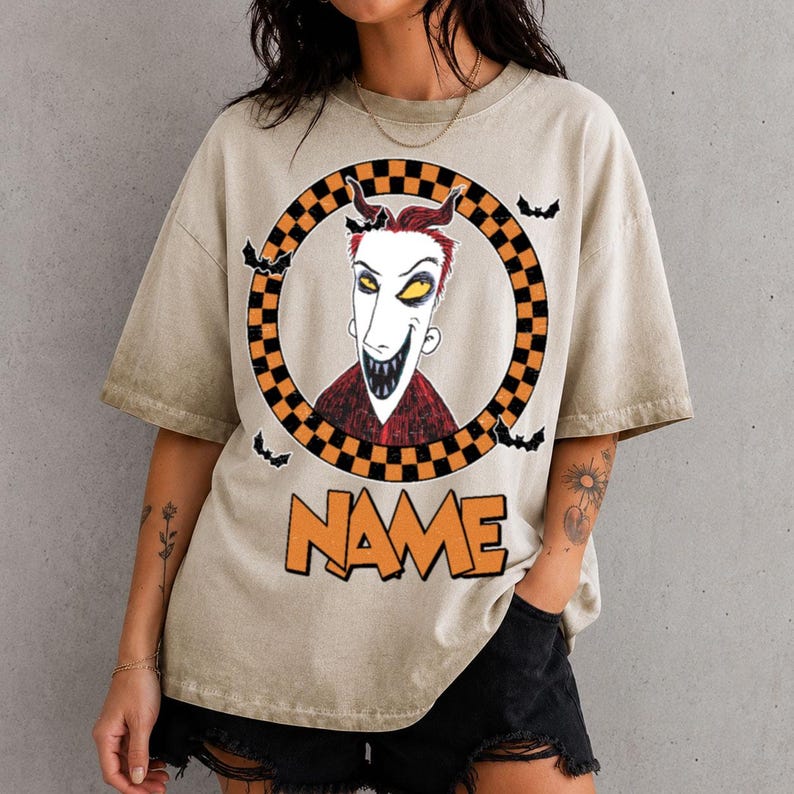 May include: A beige short-sleeve t-shirt featuring a graphic of a character with red hair, yellow eyes, and a wide grin inside a black and orange checkered circle. Small black bats surround the circle, and the word NAME is printed in orange.