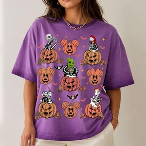 May include: A purple short-sleeved t-shirt featuring a graphic print of various characters from The Nightmare Before Christmas and Mickey Mouse-shaped pumpkins. The design includes orange jack-o'-lanterns, small bats, and yellow stars.
