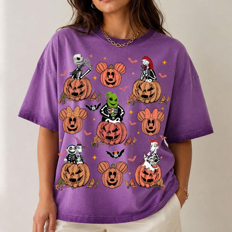 May include: A purple short-sleeved t-shirt featuring a graphic print of various characters from The Nightmare Before Christmas and Mickey Mouse-shaped pumpkins. The design includes orange jack-o'-lanterns, small bats, and yellow stars.