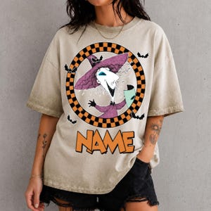 Puede incluir: Una camiseta beige de manga corta con un gr&aacute;fico de un personaje con sombrero morado dentro de un c&iacute;rculo a cuadros naranja y negro. Peque&ntilde;os murci&eacute;lagos negros rodean el c&iacute;rculo. La palabra NAME est&aacute; impresa en letras naranjas grandes.