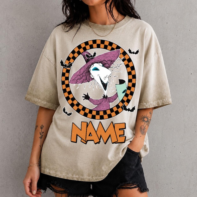 Puede incluir: Una camiseta beige de manga corta con un gr&aacute;fico de un personaje con sombrero morado dentro de un c&iacute;rculo a cuadros naranja y negro. Peque&ntilde;os murci&eacute;lagos negros rodean el c&iacute;rculo. La palabra NAME est&aacute; impresa en letras naranjas grandes.