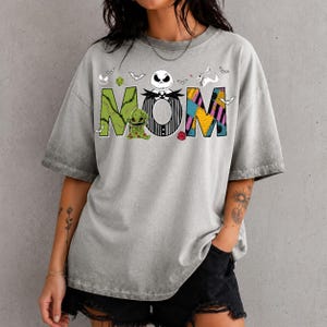 Puede incluir: Una camiseta gris con efecto lavado al &aacute;cido que presenta la palabra MOM en letras grandes y coloridas. La M es verde con un peque&ntilde;o personaje, la O es una calavera blanca con rayas negras y la M es un patr&oacute;n de parches amarillo, azul y morado con peque&ntilde;os murci&eacute;lagos.