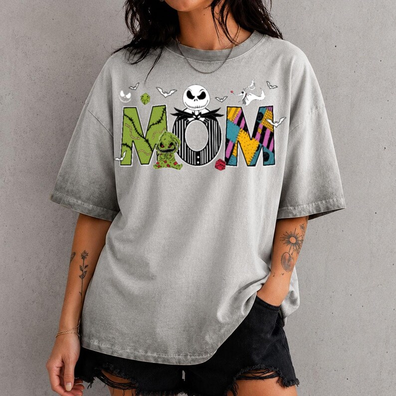 Puede incluir: Una camiseta gris con efecto lavado al &aacute;cido que presenta la palabra MOM en letras grandes y coloridas. La M es verde con un peque&ntilde;o personaje, la O es una calavera blanca con rayas negras y la M es un patr&oacute;n de parches amarillo, azul y morado con peque&ntilde;os murci&eacute;lagos.