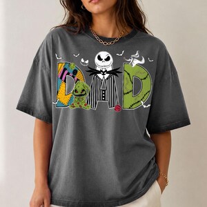 May include: A charcoal grey short-sleeve t-shirt featuring the word DAD in large letters. The D is a yellow patchwork pattern, the A is a black and white pinstripe suit with a skull head, and the D is green with stitches. Includes small bats and a ghost dog.